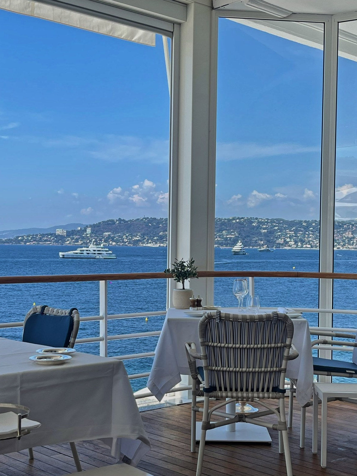 Tables set on a seaside restaurant terrace, yachts anchored in the bay beyond