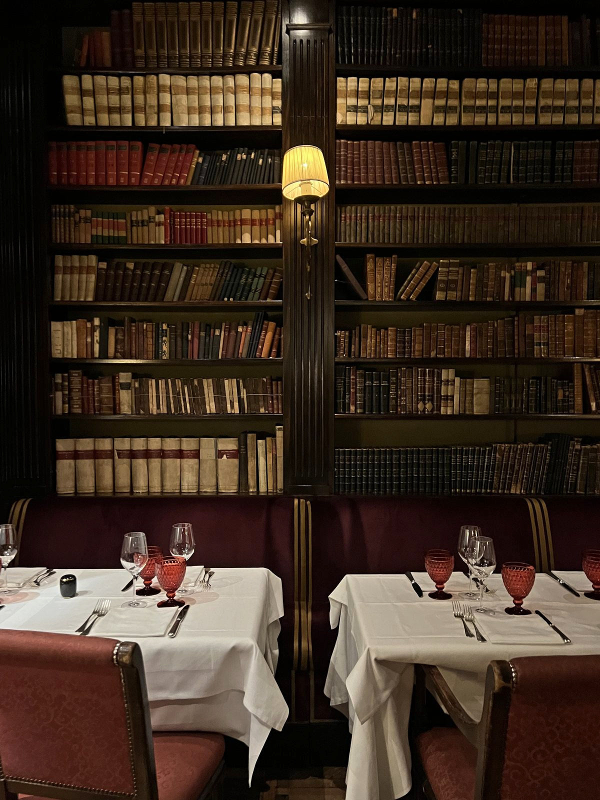 Tables laid in a restaurant lined with antique bookshelves, a sconce glowing between them