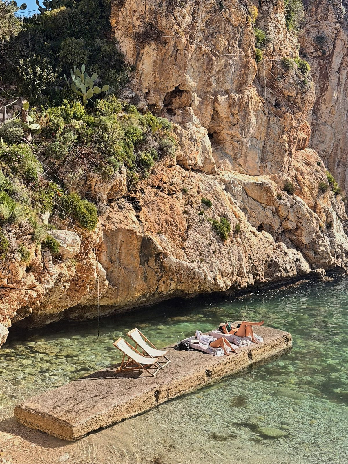 Deckchairs on a stone jetty in a clear cove below sunlit cliffs