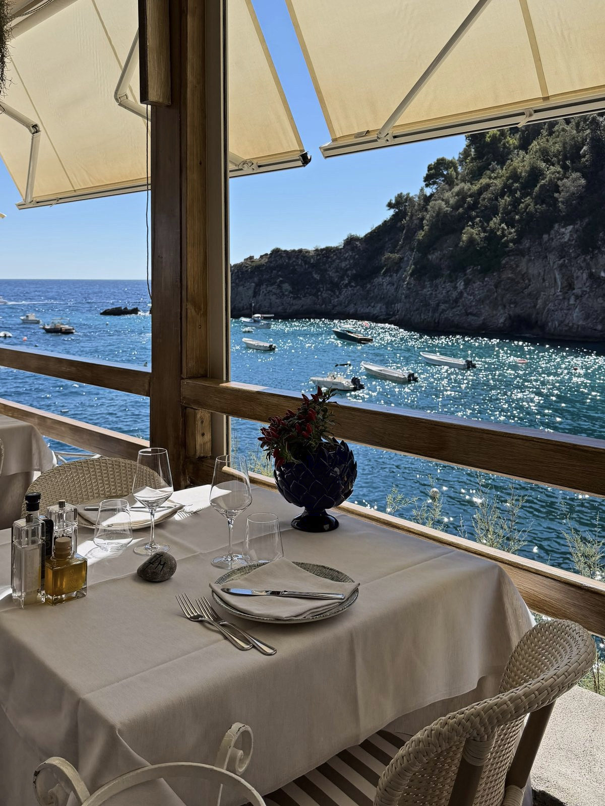 A table set on a shaded terrace above a cove, boats moored in the turquoise water below